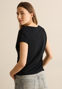 Sort kortærmet top med rund halsudskæring, der har teksturerede detaljer på skuldrene. Bæres med lysegrå jeans, der viser en afslappet pasform.