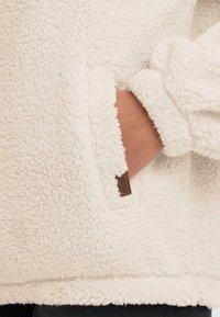 Krémová fleecová bunda s texturovaným povrchem, boční kapsou a koženou štítkovou značkou na lemu.