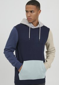 Sudadera con capucha que presenta bloques de color en navy, azul claro y beige, con una capucha gris, bolsillo delantero y puños acanalados. Textura suave.