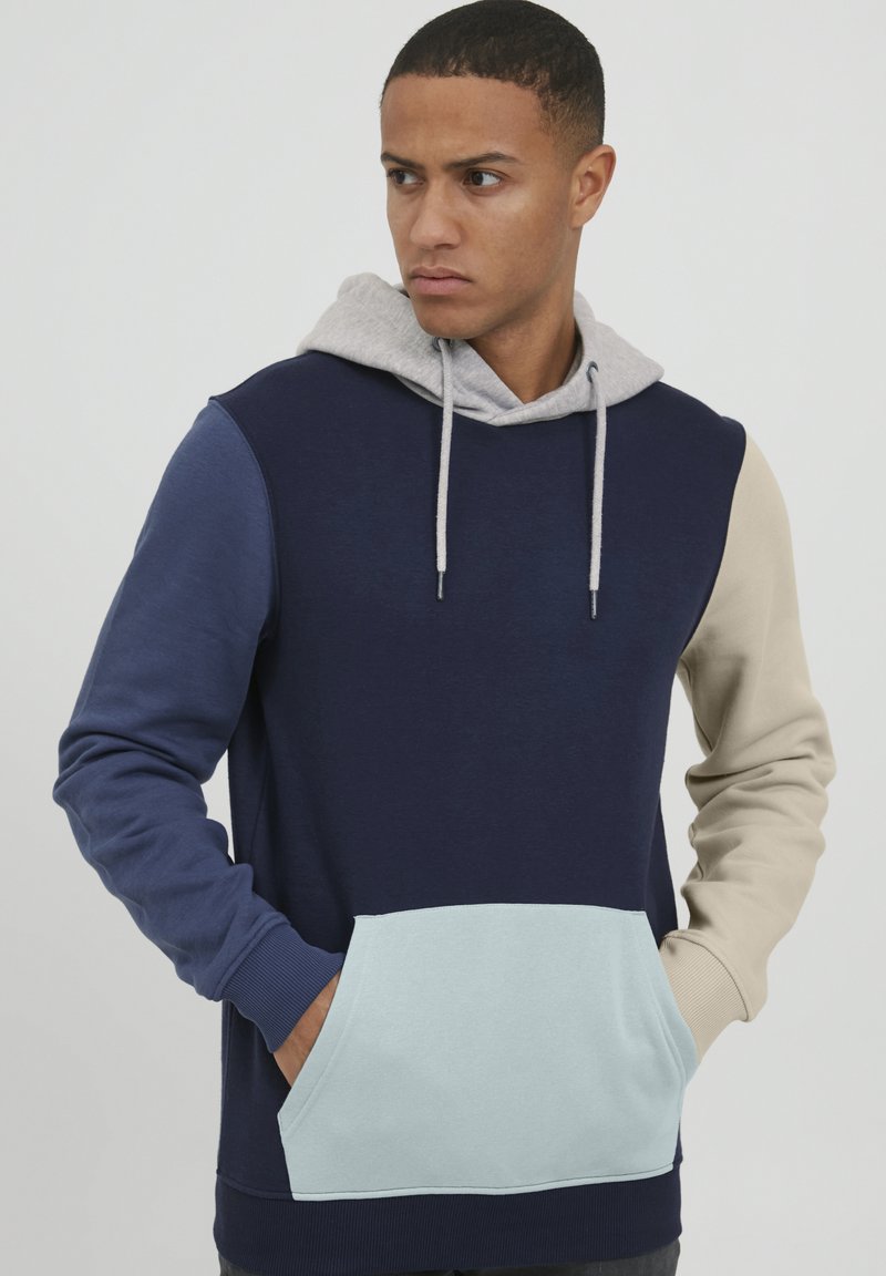 Sudadera con capucha que presenta bloques de color en navy, azul claro y beige, con una capucha gris, bolsillo delantero y puños acanalados. Textura suave.