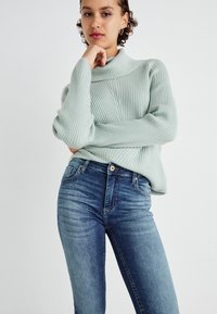 Lys grønn ribbestrikket genser med høy krage, parret med tettsittende blå denimjeans med falmede mønstre og frontlukking med knapper.