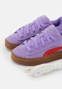 Fialové semišové tenisky s texturovanou gumovou podrážkou, vybavené šňůrkami ve stejném odstínu a červeným detailem na boku. Součástí je také dodatečná bílá šňůrka.