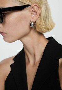 Sort blazer med skarpe revers, parret med dristige metalringe. Øreringene har en glat, skinnende overflade.