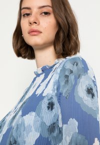 Blusa floral azul com textura pregueada, tecido leve e um decote sutilmente franzido. Flores em diferentes tons de azul.