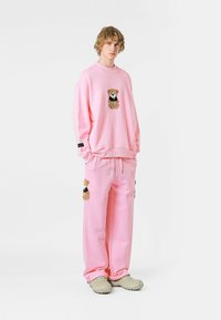 Conjunto de sudadera y pantalones rosa con parches de osito. Tejido suave, corte relajado, bolsillos laterales en los pantalones. Modelo lleva zapatos gris claro.