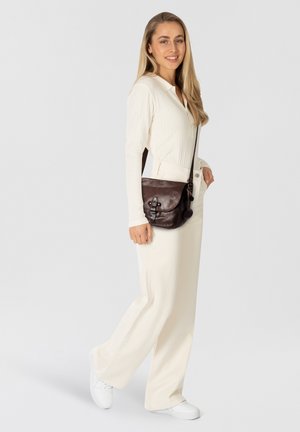 Bruine leren schoudertas met een magnetische sluiting, gebogen vorm en verstelbare schouderband, gecombineerd met een witte ribbeltop en lichte broek.