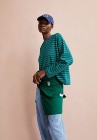 Gestreiftes langärmliges grün-blaues Hemd, kombiniert mit einem grünen Rock und hellblauen Jeans, mit einem Markenlogo und einer lässigen Baseballkappe.
