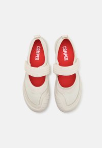 Scarpe in tessuto bianco con cinturino in velcro, lati testurizzati e una soletta rossa a contrasto con la scritta "CAMPER" stampata in bianco. Suola rotonda e imbottita.