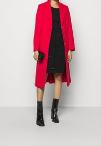 Punainen villakangastakki, jossa on muotoiltu malli, napit, kaksi taskua, yhdistetty mustan mekon ja kiiltävien mustien nilkkasaappaiden kanssa.