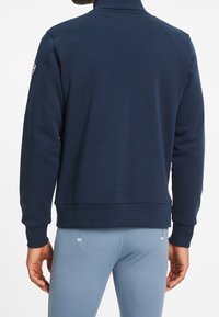 Sudadera azul marino con cuello alto, puños y dobladillo acanalados. Presenta un pequeño logo en la manga y una textura suave.