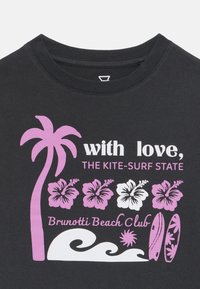 T-shirt nera con grafiche stampate in rosa e bianco di palme, fiori di ibisco, tavole da surf e testo relativo a un beach club.