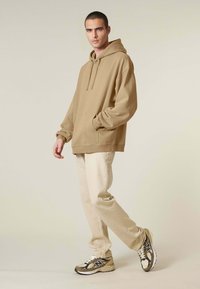 Khaki oversize mikina s kapucňou a šnúrkou, nosená s béžovými nohavicami a teniskami rôznych farieb. Hladká tkanina, základný dizajn, bočná kapsa.