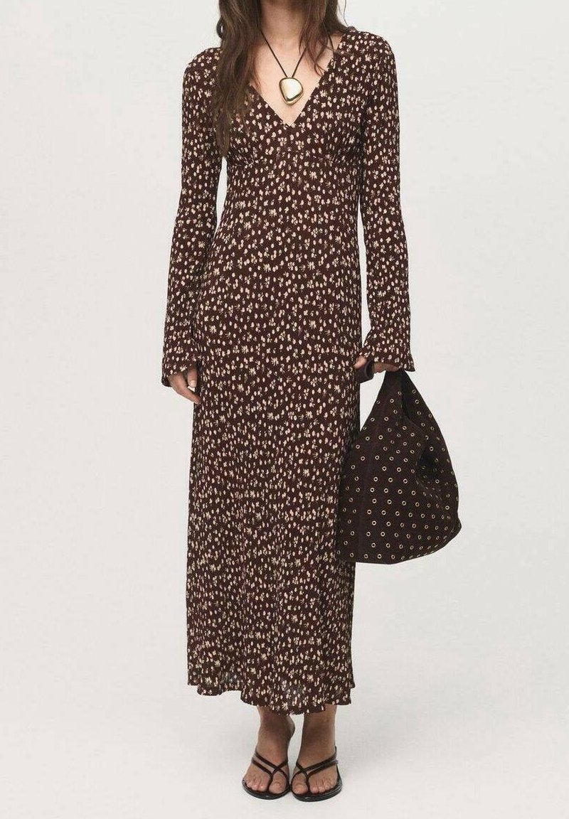 Femme en longue robe marron à fleurs tenant un grand sac fourre-tout sombre avec des motifs circulaires, portant des sandales noires à brides et un grand collier pendentif.