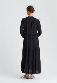 Schwarzes gestuftes Maxikleid mit langen Ärmeln, einem gerafften Taillenbereich und einem fließenden Rock. Kombiniert mit silberfarbenen Sandalen.