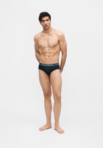 Άνδρας μοντέλο που στέκεται ξυπόλητος, φοράει μαύρα εσώρουχα Emporio Armani, με τα χέρια πίσω από την πλάτη, μπροστά σε απλό γκρίζο φόντο.