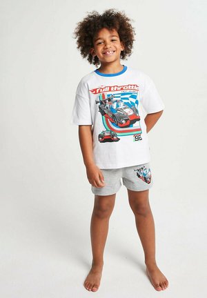 Smiling child standing barefoot wearing white Hot Wheels t-shirt with racecar graphic and gray shorts with matching car design.