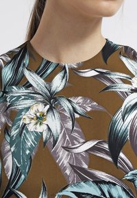 Hnědá tkanina se tropickým květinovým vzorem s velkými tyrkysovými listy a krémovými květy, zdůrazňujícími podrobné linie a textury.