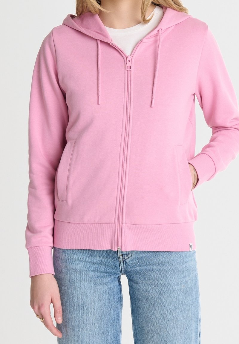 Femme portant un sweat à capuche zippé rose clair sur un t-shirt blanc avec un jean bleu clair, une main dans la poche et une main détendue sur le côté.