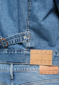 Τζιν παντελόνι σε ανοιχτό μπλε χρώμα, με δερμάτινη ετικέτα που φέρει την ένδειξη "TOMMY JEANS" και καστανές λεπτομέρειες, καθώς και μεταλλικά εξαρτήματα και ραφές.