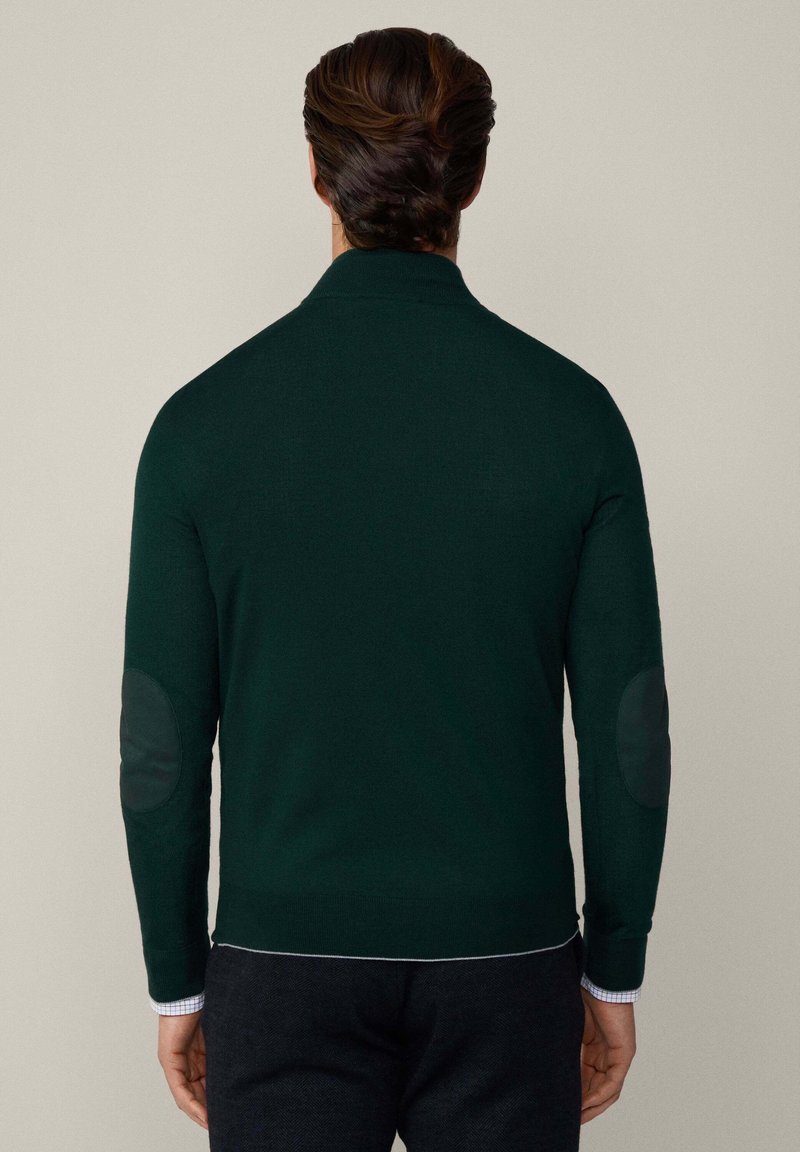 Dunkelgrüner Pullover mit hohem Kragen, tailliertem Design und strukturierten Ellenbogenpatches. Mit einem weißen Saum am unteren Rand.