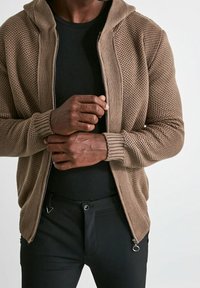 Felpa con zip lavorata a maglia di colore talpa, con pannelli testurizzati, polsini a coste e cappuccio morbido. Indossata sopra una maglietta nera e abbinata a pantaloni neri.