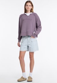 Fioletowa krótka bluza z haftowanym napisem, połączona z jasnoniebieskimi szortami dżinsowymi. Model w beżowych butach na slip-on i białych skarpetkach.