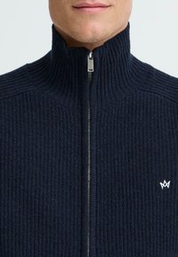Námořnický pletený svetr s vysokým límcem a plným zipem. Obsahuje malé bílé logo na hrudi a strukturovanou pletenou tkaninu.