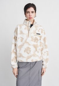 Νεαρό άτομο που φοράει μπεζ και λευκό φλις πουλόβερ με σχέδιο και λογότυπο, καθώς και γκρι cargo φούστα, που στέκεται μπροστά σε απλό ανοιχτόχρωμο φόντο.