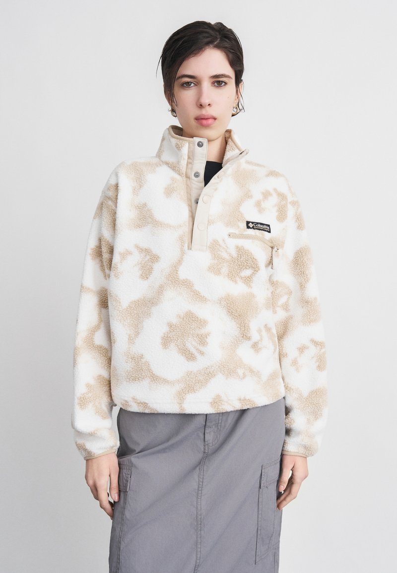 Νεαρό άτομο που φοράει μπεζ και λευκό φλις πουλόβερ με σχέδιο και λογότυπο, καθώς και γκρι cargo φούστα, που στέκεται μπροστά σε απλό ανοιχτόχρωμο φόντο.