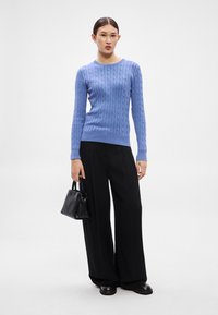 Gaiši zils džemperis ar adījumu un garām piedurknēm, kails ar melniem platu kāju biksēm un mazu melnu rokassomu. Gluda tekstūra, ikdienišķs dizains.