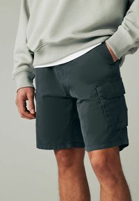 Dunkelgraue Cargo-Shorts mit aufgesetzten Taschen und entspannter Passform, aus leichtem Material gefertigt. Getragen mit einem hellgrauen Sweatshirt.