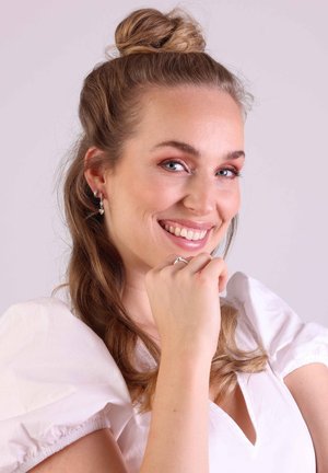 Des boucles d'oreilles en argent pendent avec des accents de pierres précieuses transparentes. Le modèle porte un haut blanc à manches bouffantes, mettant en valeur de longs cheveux ondulés.