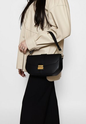 Donna con giacca beige e gonna nera che tiene una borsa a mano in pelle nera con fibbia dorata e tracolla.