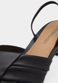 Gros plan sur une sandale en cuir noir avec une lanière matelassée croisée et une fine lanière à la cheville sur une semelle beige.