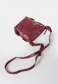 Bordeaux leren schoudertas met een zachte textuur, ritssluiting, verstelbare schouderband en decoratieve hardwareaccenten. Compact rechthoekig van vorm.