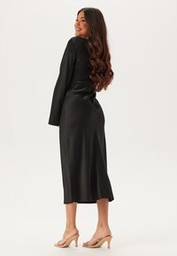 Zwarte satijnen jurk met lange mouwen en een aansluitende silhouette, die tot halverwege de kuit reikt. Heeft een gladde textuur en een lichte glans.