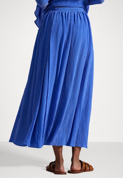 Lange blauwe rok van gestructureerde stof, met een geplooide tailleband en zijslits, gecombineerd met bruin kleurige sandalen.