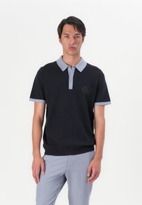 Polo em malha preta com gola cinza e detalhes nas mangas, apresentando um padrão texturizado discreto e um logótipo redondo no lado esquerdo.