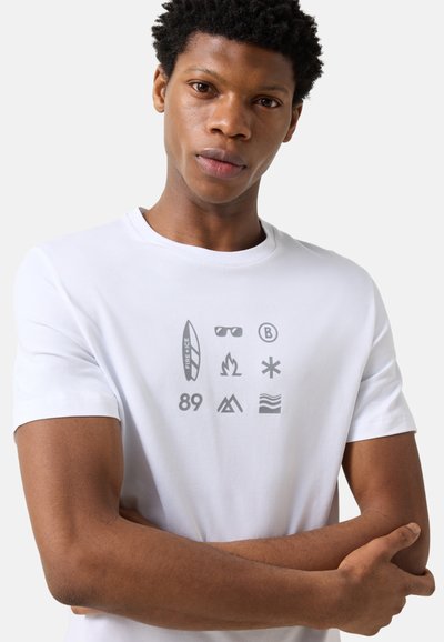 Homme portant un T-shirt blanc avec des icônes grises incluant une planche de surf, des lunettes de soleil, du feu, un astérisque, le numéro 89, une montagne et des vagues, les bras croisés.
