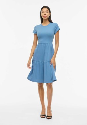Kobieta stoi, ubrana w niebieską sukienkę z krótkim rękawem, sięgającą do kolan, z marszczonym dołem, w połączeniu z czarnymi, odkrytymi czubkami butów na białym tle.