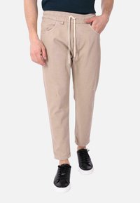 Pantaloni jogger beige con vita elastica e cordoncino. Presentano tasche frontali e piccoli dettagli di usura. Abbinati a sneakers nere.