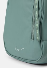 Nike ryggsäck i dämpad grön, tillverkad av texturerad tyg. Har en dragkedja med varumärkesband och en vit logotyp på botten.