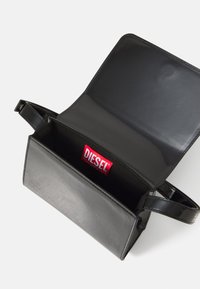 Borsa a tracolla in ecopelle nera con una texture liscia e una chiusura a pattina. L'interno presenta un'etichetta rossa con il logo "DIESEL". Forma rettangolare compatta.