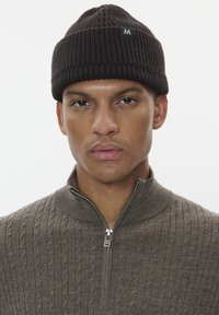 Brown ribbed knit beanie with a small black tag. Worn with a gray zip-up sweater featuring a textured pattern. Close-up of the face.