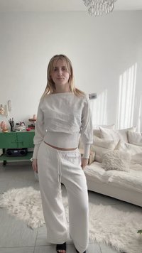 Νέα γυναίκα με λευκό πουλόβερ και παντελόνι στέκεται σε ένα φωτεινό σαλόνι με πράσινη κασέλα, καναπέ με μαξιλάρια και ένα χνουδωτό χαλί.