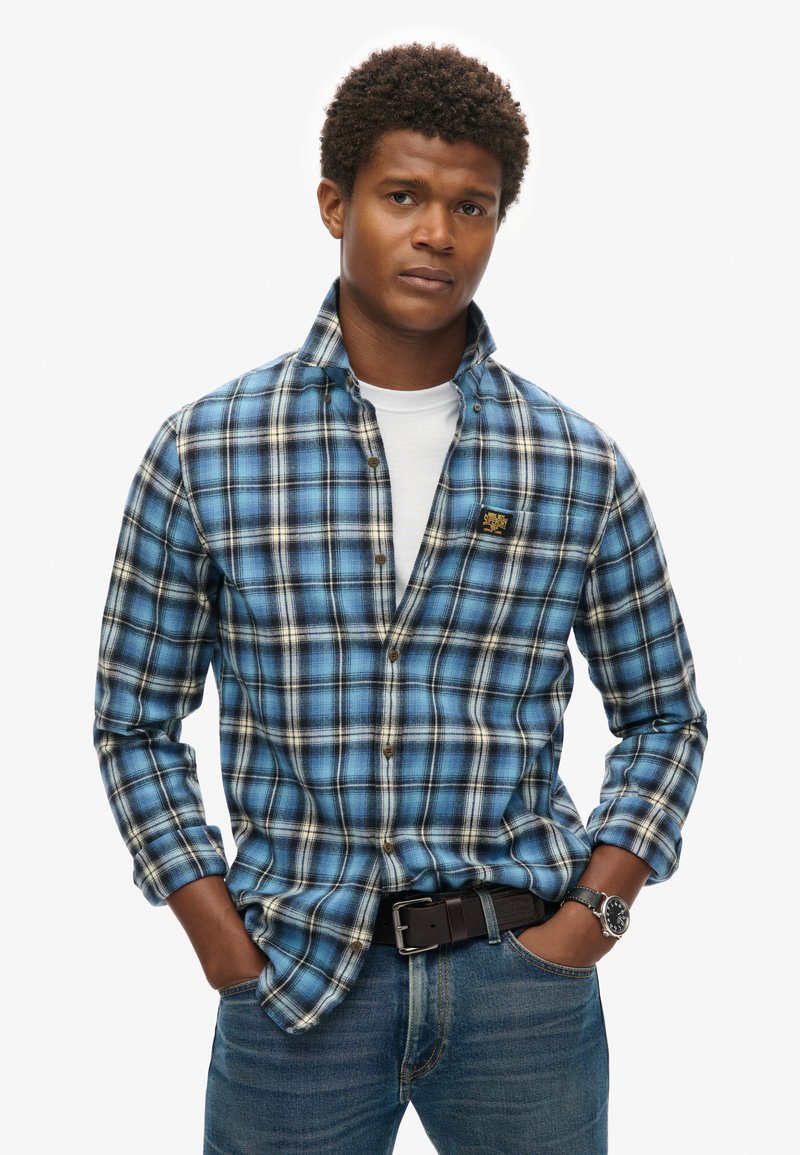 Camicia in flanella a quadri blu e neri con colletto button-down, caratterizzata da dettagli di un beige chiaro. Indossata sopra una camicia bianca, abbinata a jeans e una cintura nera.