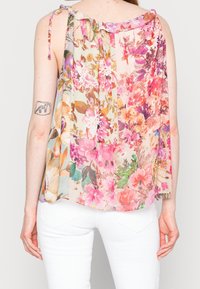 Blusa floral transparente con un escote con volantes, en tonos rosa, púrpura y crema, con tirantes atados y un corte holgado. Combinada con pantalones blancos.
