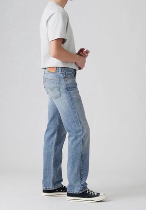 Person iført lyseblå jeans med lige ben, en lysegrå kortærmet skjorte og sorte lave sneakers, stående med hænderne foldet.