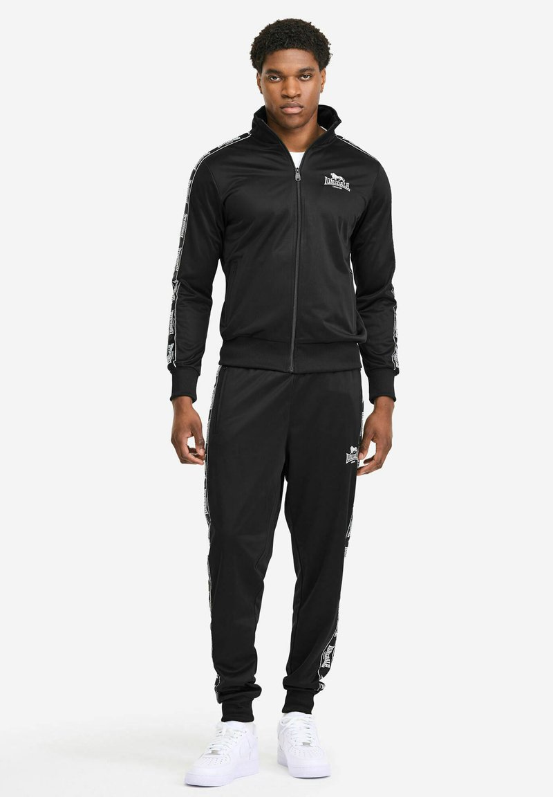 Black tracksuit made of smooth fabric, featuring a zip-up jacket with a high collar and matching pants. Includes white logo accents and stripes.