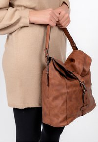 Sac en cuir marron à finition texturée, avec une large ouverture, une sangle réglable et un rabat de fermeture, tenu contre un fond clair.